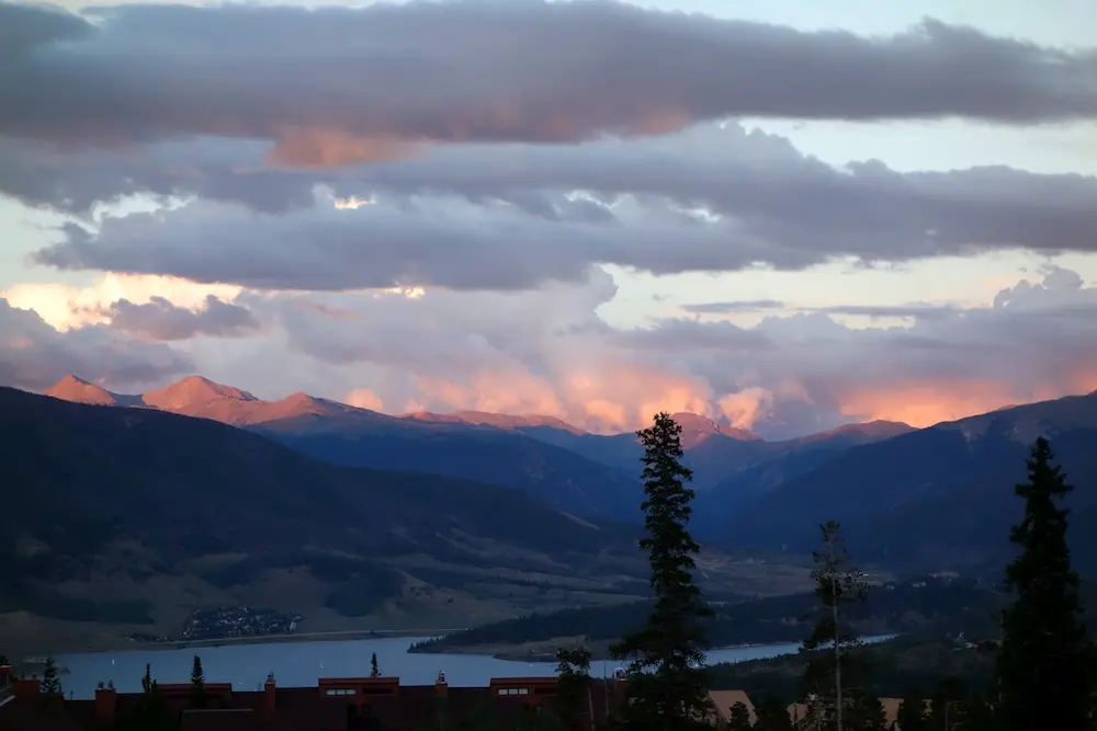 Sunset light hits mountain peaks under a cloudy sky, with dark hills and scattered buildings in the foreground and a lake in the distance.