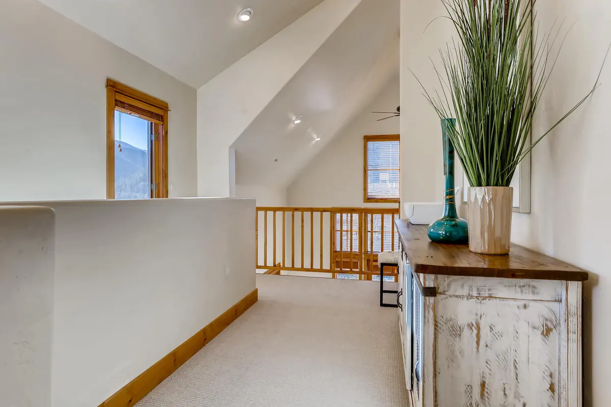 Upstairs hallway with beige carpet, white walls, wood trim, a rustic sideboard with tall green plants, and a wooden railing overlooking a bright loft area.
