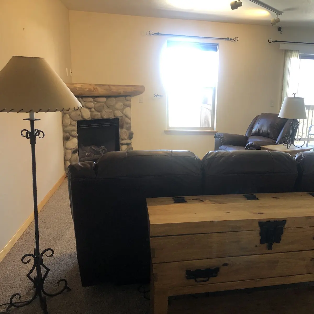 A cozy living room with a stone fireplace, brown leather sofa and chairs, wooden chest, floor lamp, and natural light coming through windows with blinds.