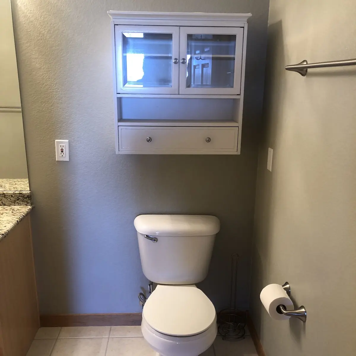 White toilet beneath a wall-mounted cabinet with glass doors. Toilet paper holder and empty towel rack are on the right wall; granite countertop and light switch are on the left. The walls are painted light beige.