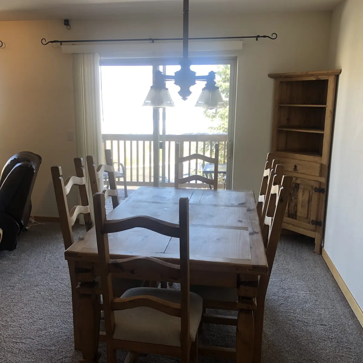 A wooden dining table with six chairs sits in a carpeted room. A wooden hutch is in the corner, and glass doors lead to a balcony with a view outside. A hanging light fixture is above the table.