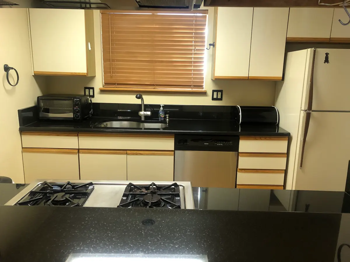 A modern kitchen with cream cabinets, black countertops, a white fridge, dishwasher, toaster oven, and a double gas stove. A window with wooden blinds is above the sink.