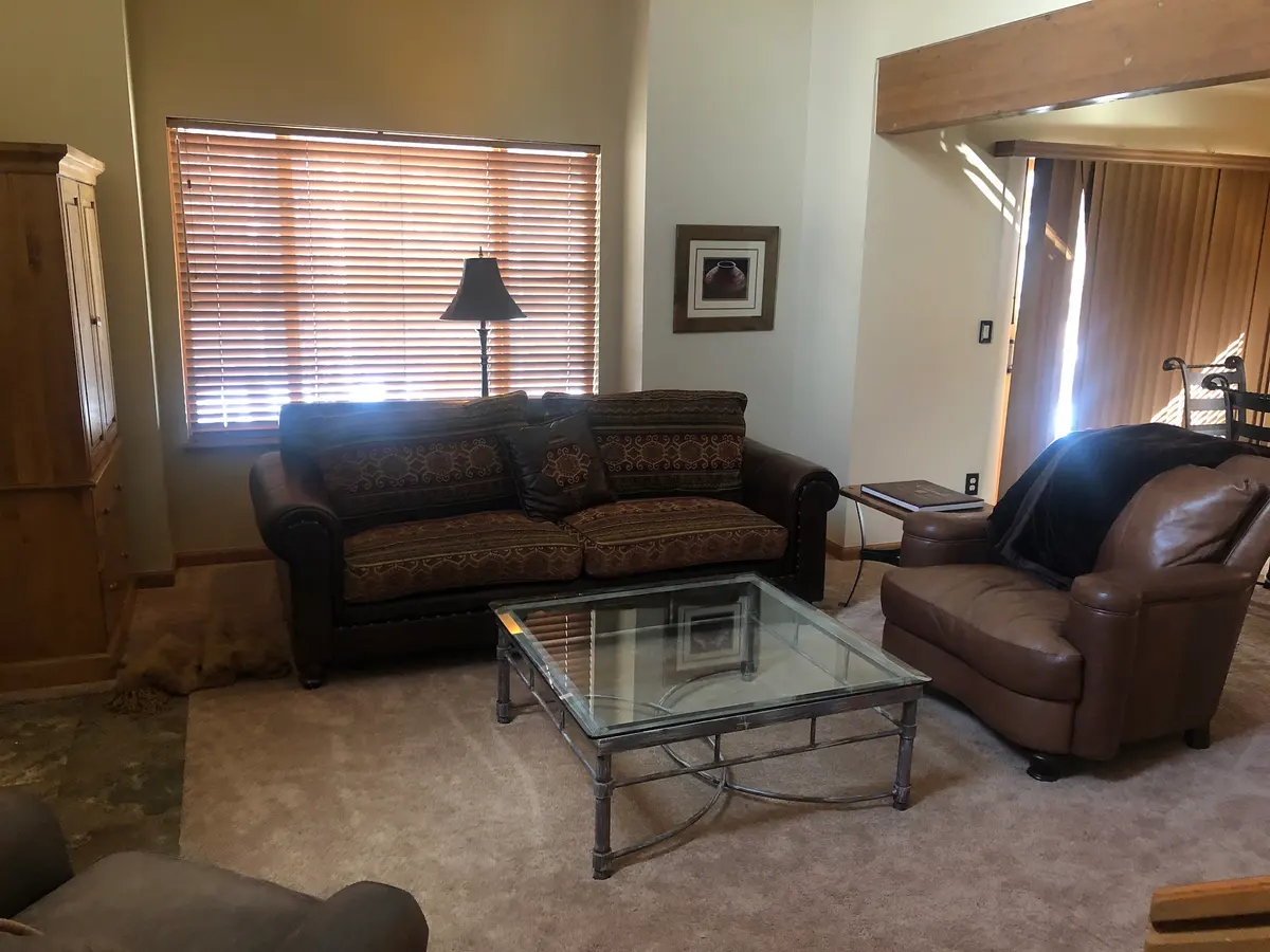 Living room with a patterned sofa, leather armchair, glass coffee table, floor lamp, wooden blinds, framed wall art, and neutral-colored carpet.