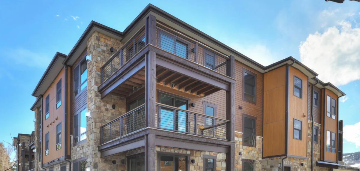 Modern three-story apartment building with stone and wood siding, large glass windows, and spacious balconies with metal railings, under a bright blue sky with clouds.