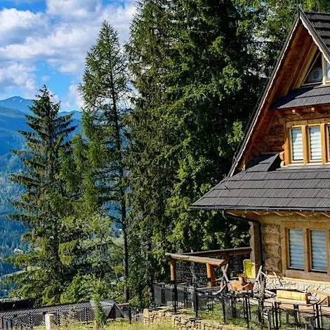 A wooden cabin with a dark roof sits on a hillside, surrounded by tall pine trees and mountains under a blue sky with clouds.