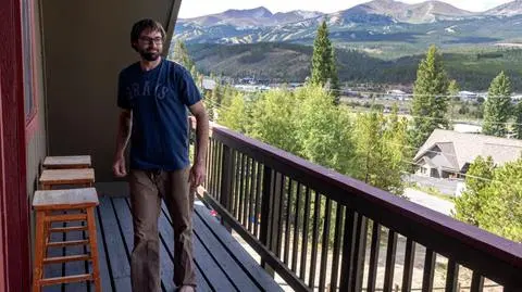 A man with glasses and a beard stands on a balcony with wooden stools, overlooking a scenic landscape of trees, houses, and mountains in the background.