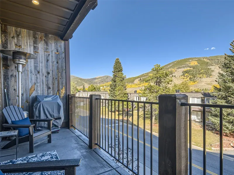 A mountain view from a balcony with a grill, patio chairs, and a small table, overlooking a road, trees, and distant hills under a clear blue sky.
