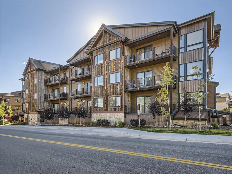 A modern, three-story apartment building with wood paneling, large windows, and balconies, pictured on a sunny day with clear skies and a road in front.
