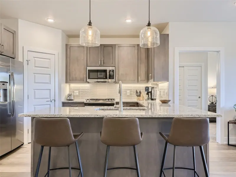 Modern kitchen with gray cabinets, stainless steel appliances, a central island with granite countertop, three brown barstools, pendant lights, and light wood floors. A door and a small glimpse of another room are visible.