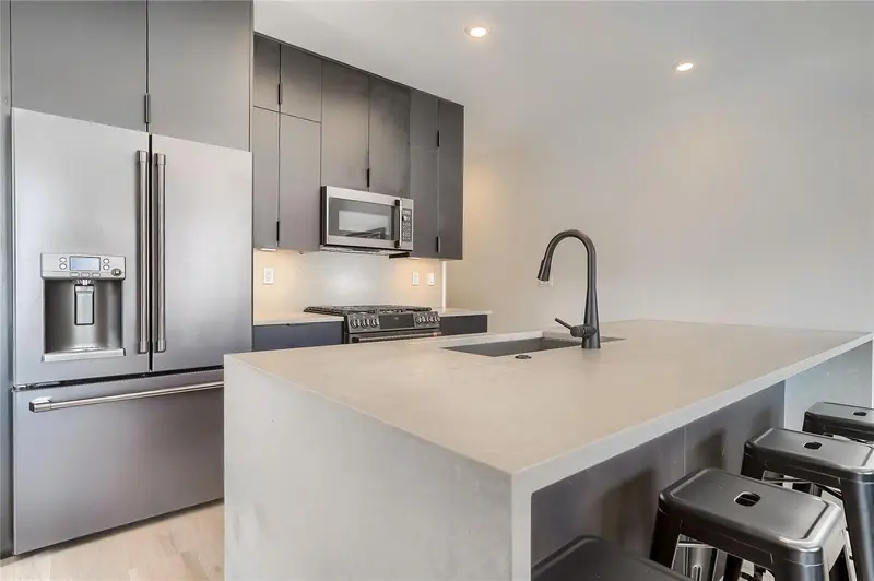 Modern kitchen with gray cabinets, stainless steel appliances, a built-in microwave, and a large island featuring a sink and black barstools. Bright lighting and minimalist design create a clean, contemporary look.