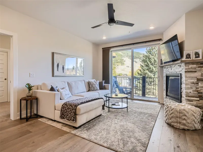 Modern living room with a beige sectional sofa, round glass coffee table, wall-mounted art, fireplace with stone surround, and large window offering a scenic mountain view. Ceiling fan and neutral decor complete the cozy space.