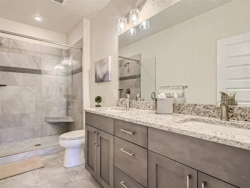 Modern bathroom with double sink vanity, granite countertop, large mirror, walk-in glass shower, gray cabinets, and neutral tile flooring. Towel and tissue box are on the counter, and framed art hangs on the wall.