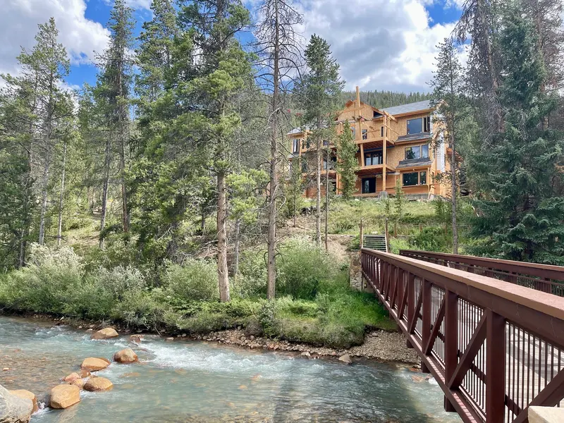 A wooden house sits among tall pine trees near a river, with a metal footbridge crossing the water in the foreground and mountains and clouds in the background.