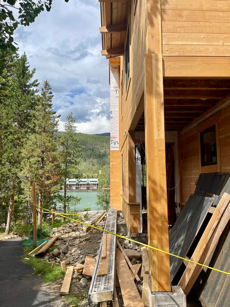 A wooden house under construction near a lake, with trees, mountains, and a dock in the background. Construction materials and yellow caution tape are visible in the foreground.