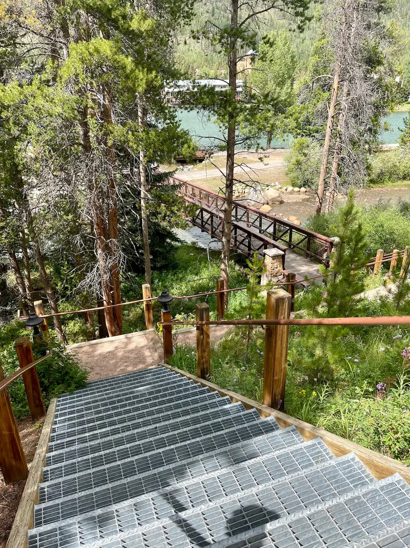 Metal stairs descend through a forest to a footbridge over a river, with trees, greenery, and a building visible in the background under bright sunlight.