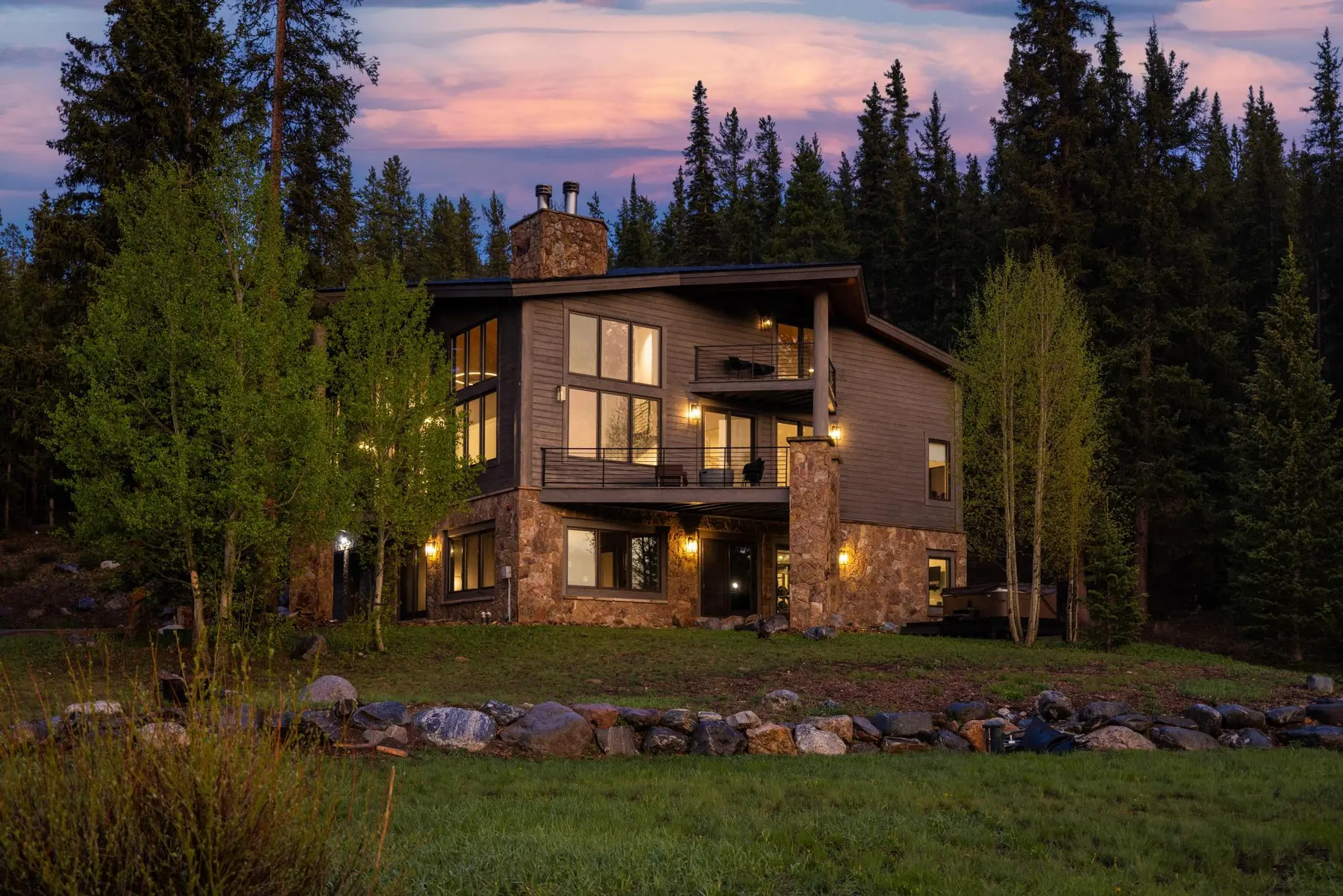 A modern mountain home with stone and wood exterior lit at dusk among evergreens
