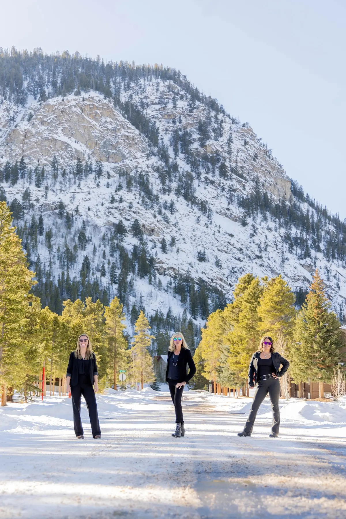 Candice De, Amy Nakos, and Jennifer Luberto standing among evergreens below a snowy peak in Frisco