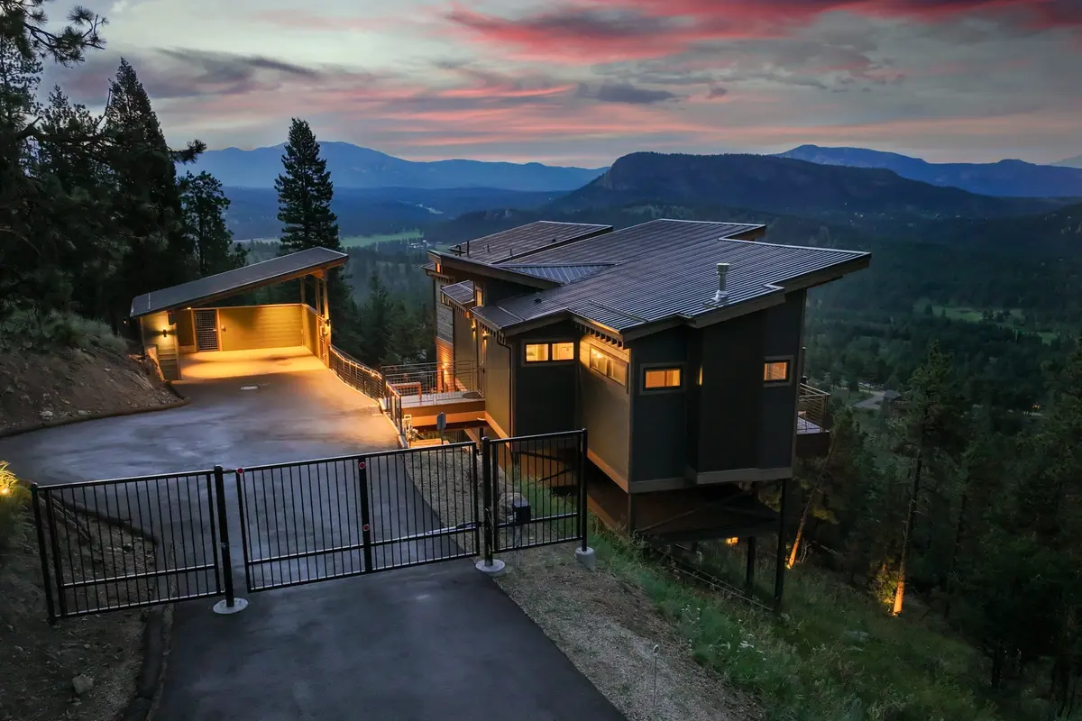 Modern house on a hillside at sunset, with a gated driveway, surrounding trees, mountains in the distance, and warm exterior lights illuminating the property.