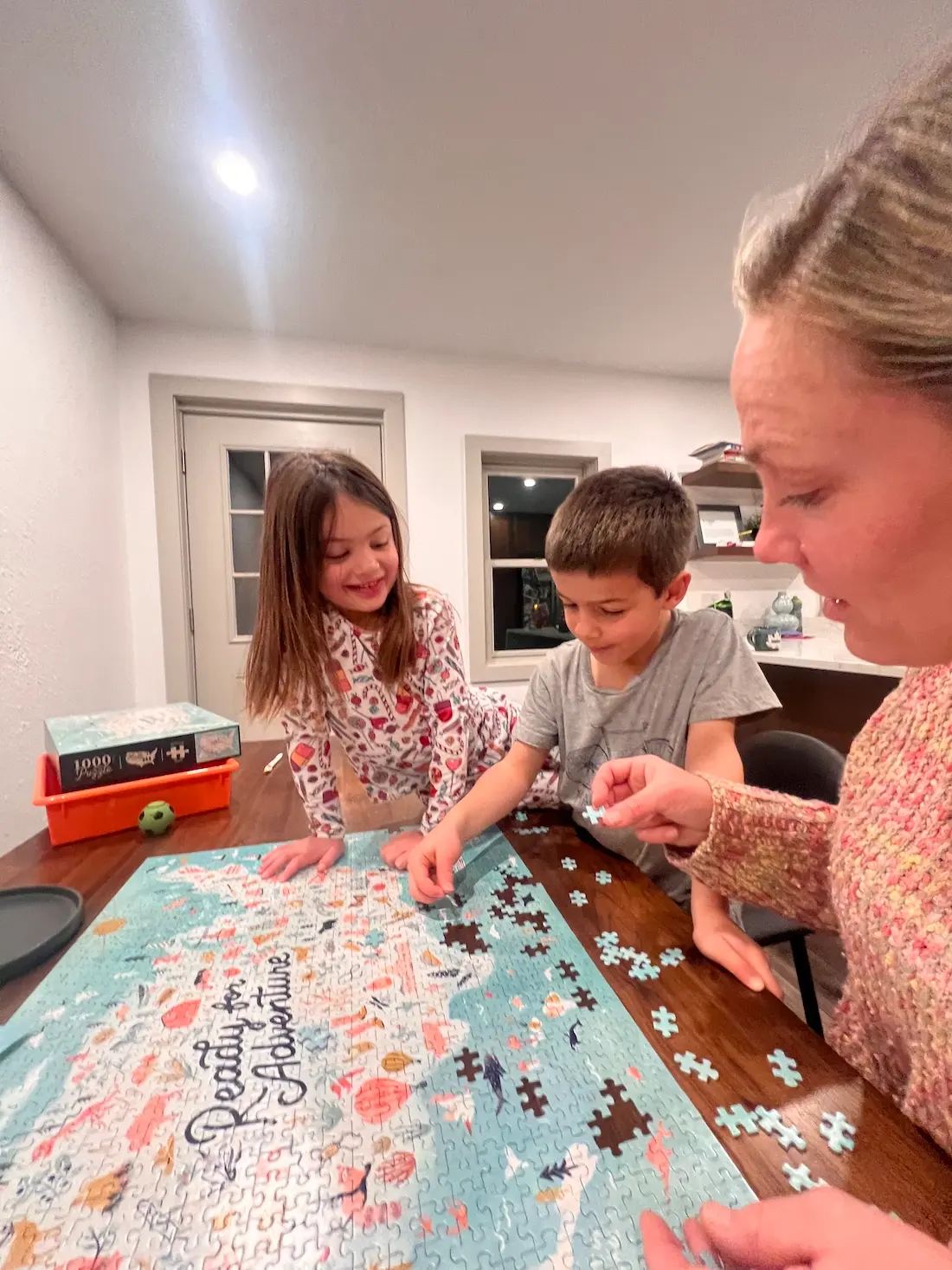 Three people sit at a table assembling a large jigsaw puzzle. A young girl and boy work together, smiling, while an adult helps. Puzzle box and extra pieces are also on the table. The setting appears to be a cozy, well-lit room.