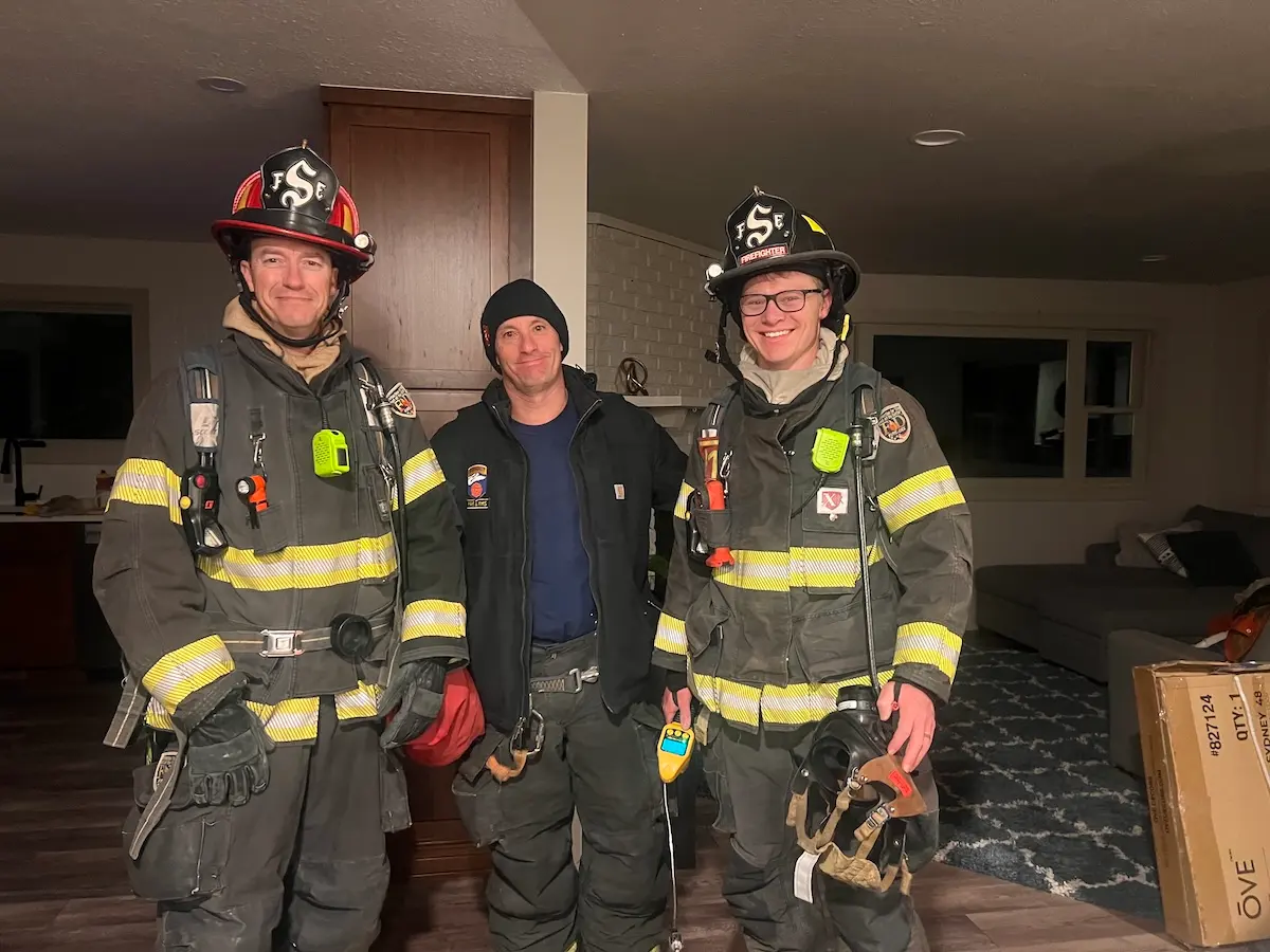 Three firefighters, two in full gear and one in a black jacket and beanie, stand and smile indoors. The room has wooden floors, a couch, a rug, and a cardboard box in the corner.
