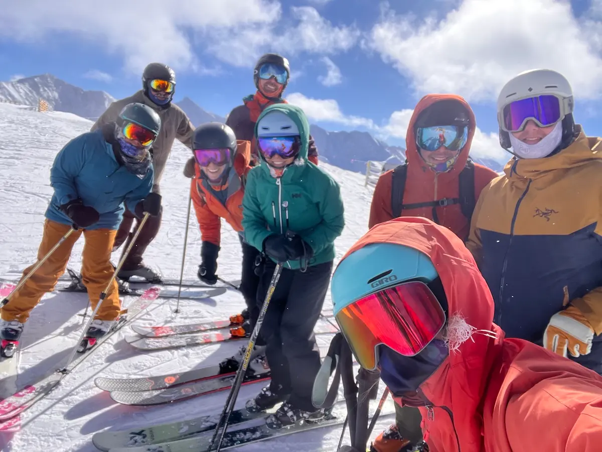 A group of eight people in colorful ski gear pose together on a snowy mountain slope, with skis and poles, under a sunny, partly cloudy sky.