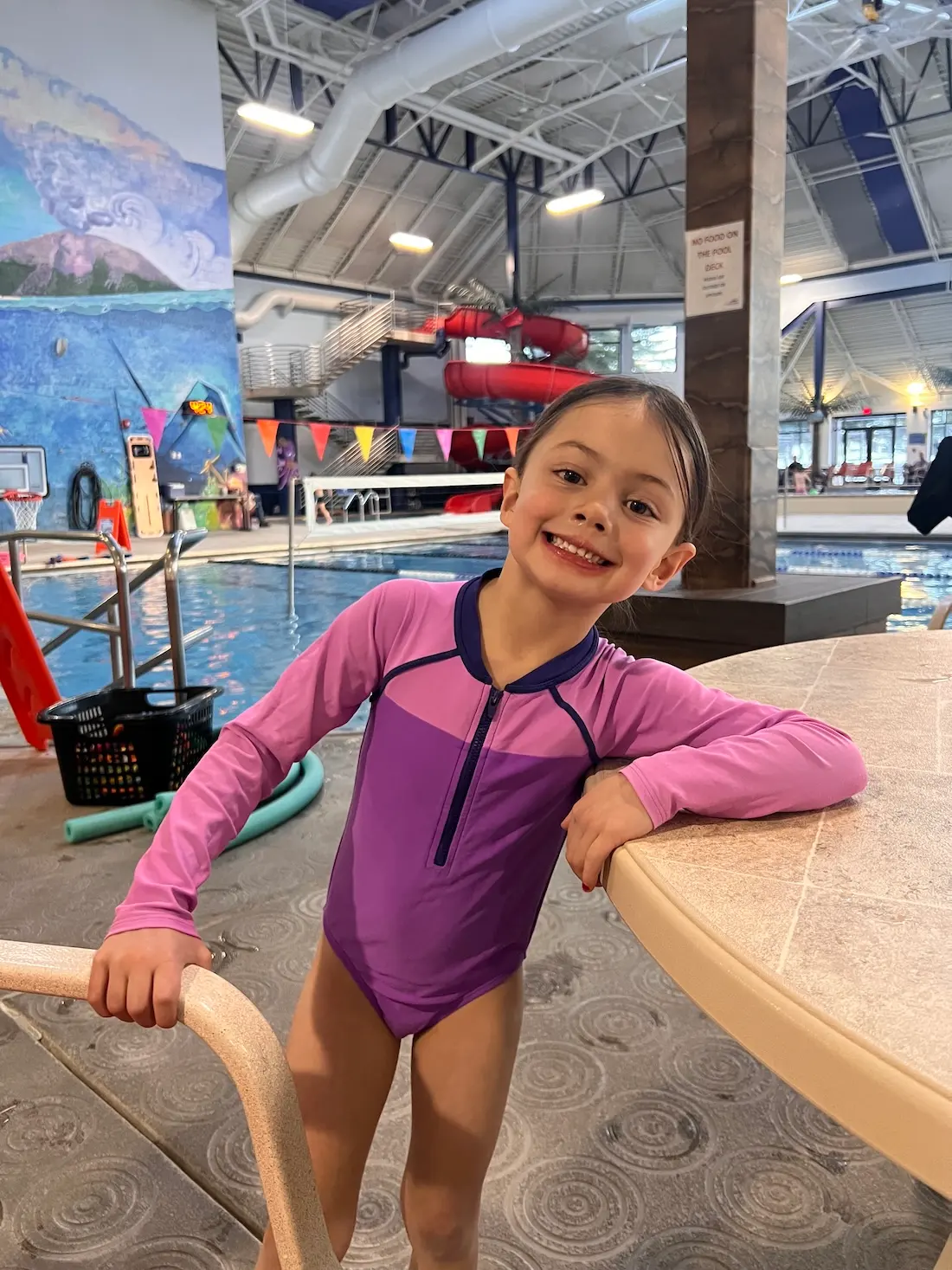 A smiling young child in a purple swim outfit stands by an indoor pool, holding a pool ladder and leaning on a table. The pool and colorful mural are visible in the background.