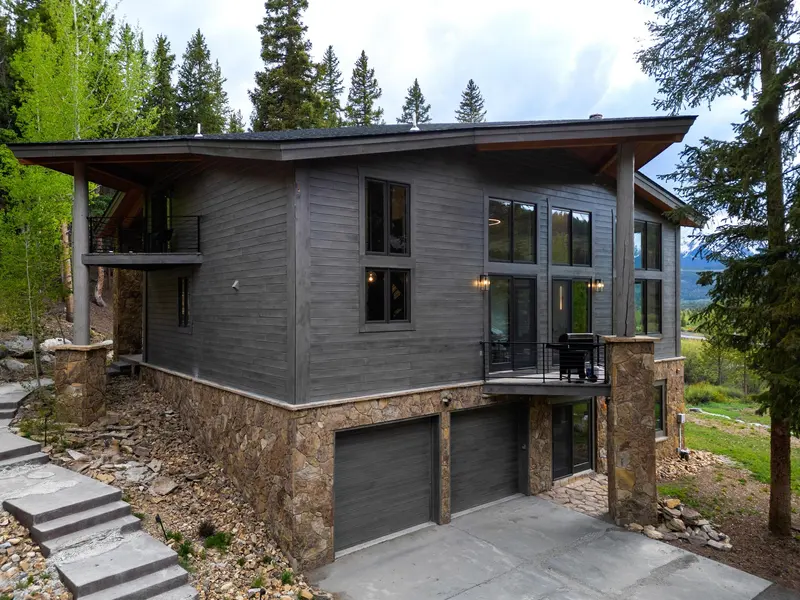 Modern two-story house with large windows, a stone and wood exterior, and a double garage, surrounded by trees and a stone pathway.
