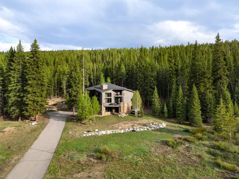 A modern house with large windows sits surrounded by dense green pine trees and a driveway, set in a forested area under a partly cloudy sky.