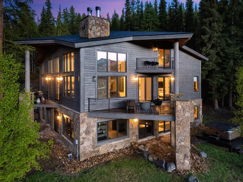 Modern mountain home with large windows, stone accents, and warm interior lights, surrounded by trees at dusk. The house has multiple balconies and a hot tub on the ground level.