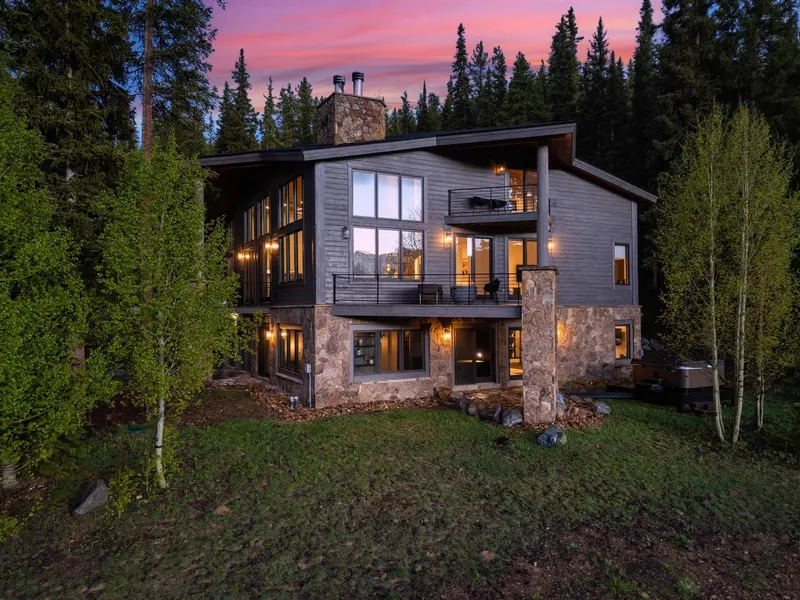 Modern two-story house with large windows, stone and wood exterior, glowing warm lights, surrounded by tall trees and greenery at sunset with a pink and purple sky.