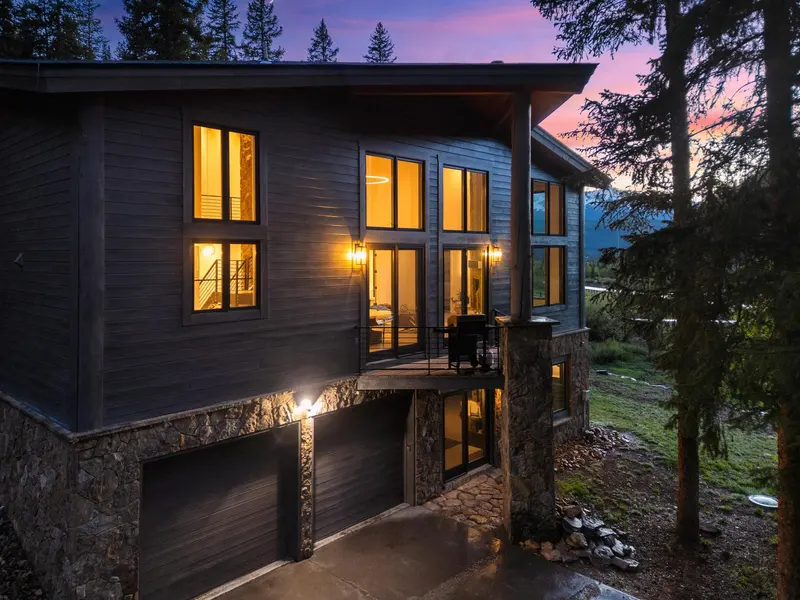 Modern two-story house with large windows, stone and wood exterior, double garage, and outdoor lights illuminated at dusk, surrounded by trees and nature.