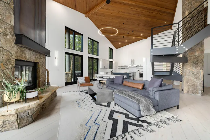 Modern living room with high wooden ceiling, gray sectional sofa, cozy rug, spiral staircase, stone fireplace, and large windows with forest views. Dining and kitchen areas are visible in the background.