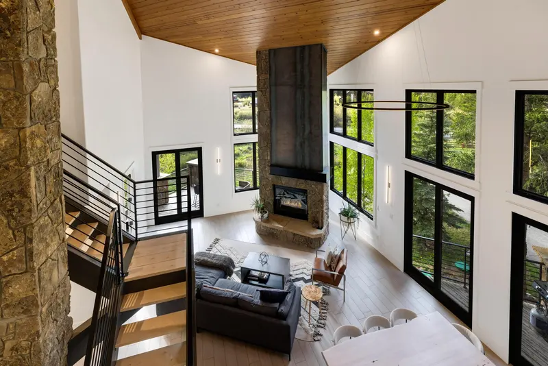 Modern living room with large windows, gray sectional sofa, black fireplace with stone base, light wood floors, and black metal staircase. Tall ceiling with wood paneling adds warmth. Greenery is visible outside through the windows.