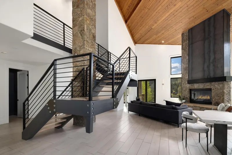 Modern living room with high wood-paneled ceiling, stone and metal staircase, black sofa, fireplace, large windows, and light wood floors. Contemporary, open-concept design with natural light.