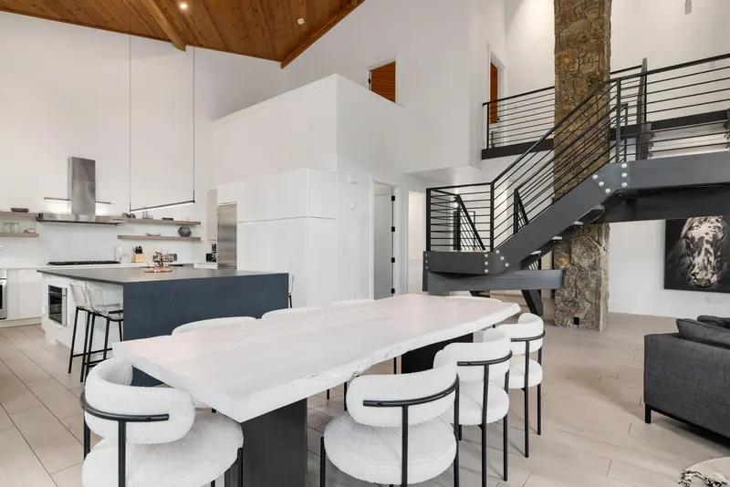 Modern open-concept kitchen and dining area with a white table, six chairs, dark staircase with metal railings, stone accent wall, and high wood-paneled ceiling.