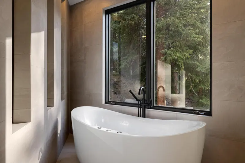 A modern bathroom features a freestanding white bathtub below a large window with a view of tall green trees outside. The walls are beige with recessed shelves.