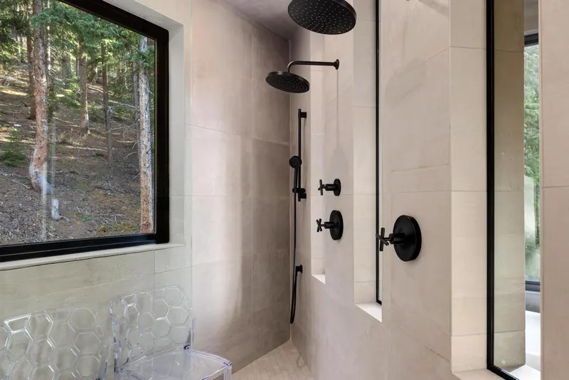 Modern shower with dual black showerheads, large beige tiles, and two windows showing a forest view. A clear, hexagon-patterned chair is visible beside the shower.