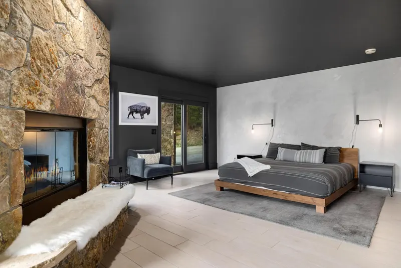 Modern bedroom with a stone fireplace, gray walls and ceiling, a large bed with striped bedding, wall-mounted lights, a plush chair, and glass doors opening to an outdoor view.