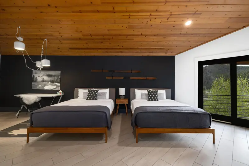 Modern bedroom with two double beds, black accent wall, wooden ceiling, and large window showing a green outdoor view. Desk with chair and wall art on the left, minimalist decor.