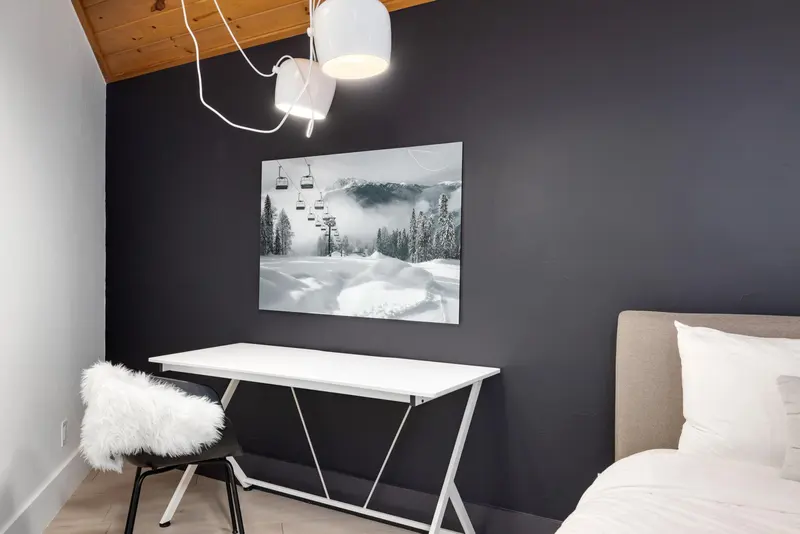 Minimalist room with a white desk, black chair with a fluffy cushion, a bed, modern ceiling light, and a wall art of snowy mountains with ski lifts. The wall is painted dark, and the ceiling is wood-paneled.