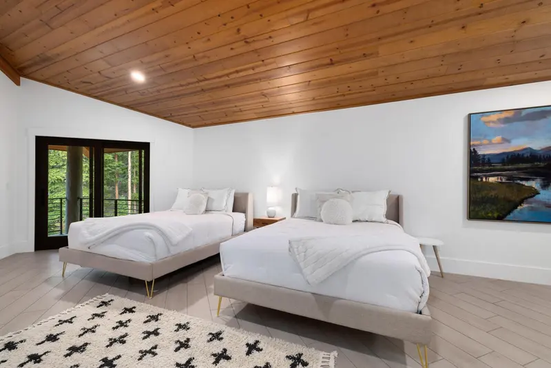 A bright bedroom with two double beds, white bedding, a wooden ceiling, a large window showing trees, a patterned rug, a nightstand with a lamp, and a landscape painting on the wall.