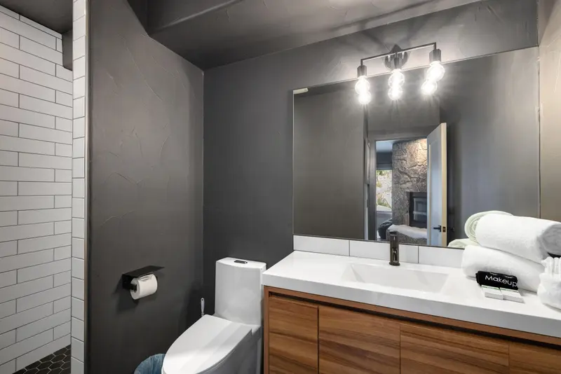 Modern bathroom with dark gray walls, a white toilet, wooden vanity with white countertop, large mirror, and three light bulbs above. A shower with white subway tiles is partially visible on the left. Towels and soap are on the counter.