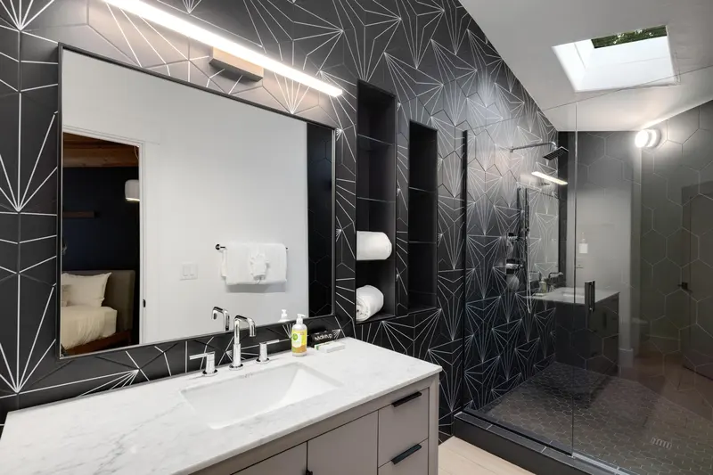 Modern bathroom with geometric black tile walls, a large mirror above a white sink, towel shelves, and a glass-enclosed shower area. A skylight provides natural light. Bedroom partially visible through an open door.