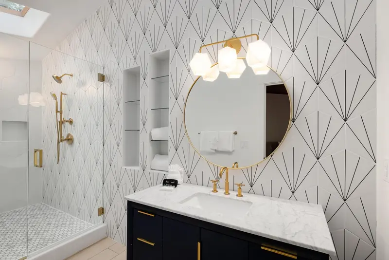 Modern bathroom with geometric white tile, gold fixtures, round mirror, marble countertop, dark vanity, and glass shower enclosure. Wall light above the mirror is on. Towels are neatly placed on a built-in shelf.