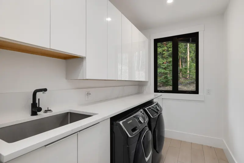 Modern laundry room with white cabinets, a black faucet over a stainless steel sink, a washer and dryer set, light wood floor, and a window showing trees outside.
