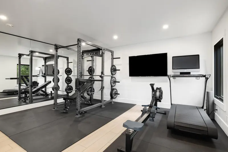 Modern home gym with a power rack, weight plates, rowing machine, treadmill, wall mirrors, and mounted screens. The room has white walls, black flooring, and large windows for natural light.