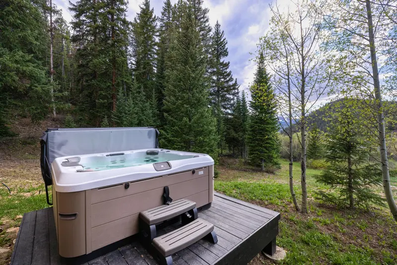 A hot tub sits on a wooden deck surrounded by green grass, trees, and forested hills under a cloudy sky.