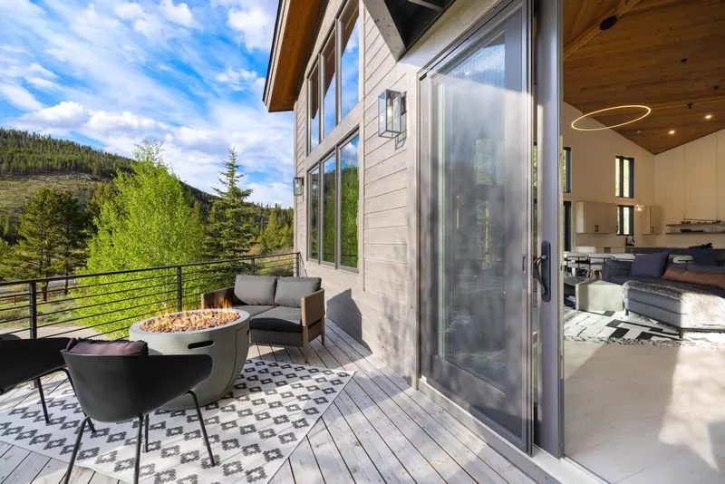 A modern mountain homes deck with a fire pit, chairs, and scenic views of trees and hills. Large windows and a glass door connect the deck to the stylish, open indoor living area.