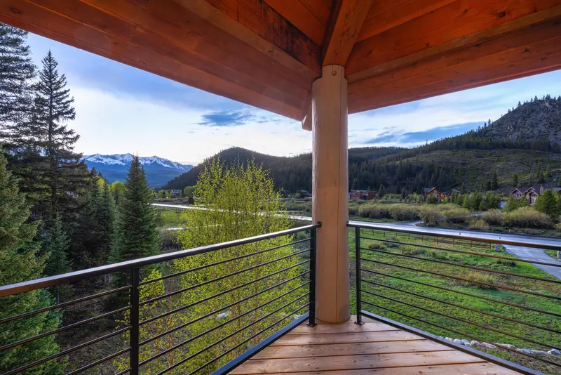 View from a wooden balcony overlooking a river, green trees, and distant mountains, partially covered in snow, under a partly cloudy sky at sunset.