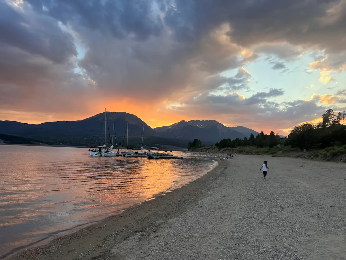 A serene lake at sunset with sailboats docked along the shore, mountains in the background, colorful clouds, and a person walking on a sandy beach.